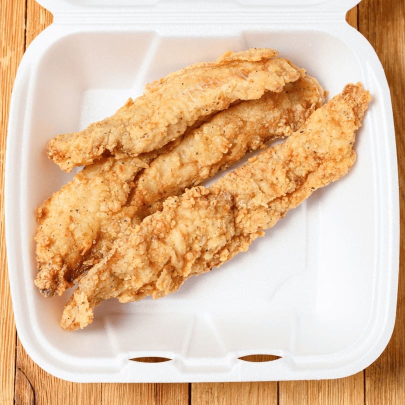Three golden-brown fried chicken tenders in a white styrofoam takeout container on a wooden surface.