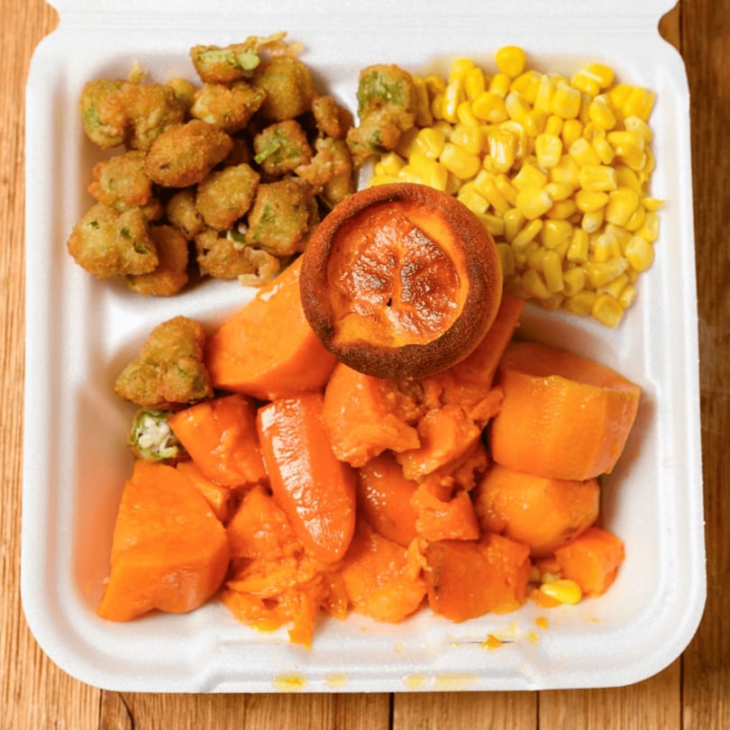 A takeout container with fried okra, corn, candied yams, and a baked peach.