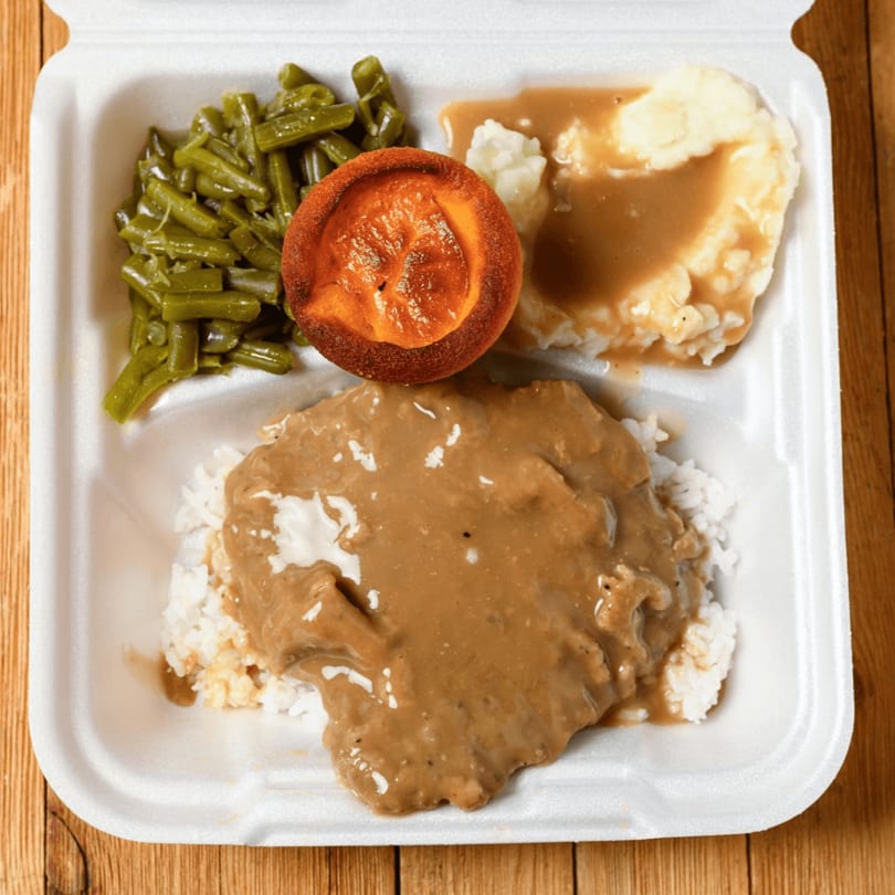 A takeout container with Salisbury steak, rice, mashed potatoes, gravy, green beans, and a roll.
